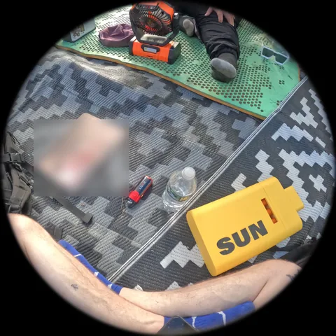 Through the camera: a camp table on a patterned mat, a yellow SUN box, crossed arms at the bottom. A screen on the mat is blurred.