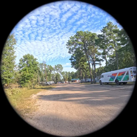 Through the camera: a sand road through the festival grounds under a rippled sky, a painted bus parked on the right.
